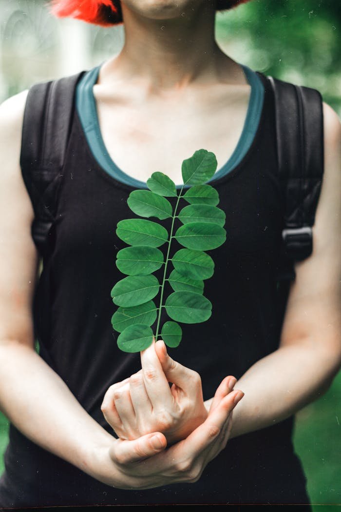 Person holding a moringa leaf while wearing a black tank top outdoors, emphasizing nature's beauty.